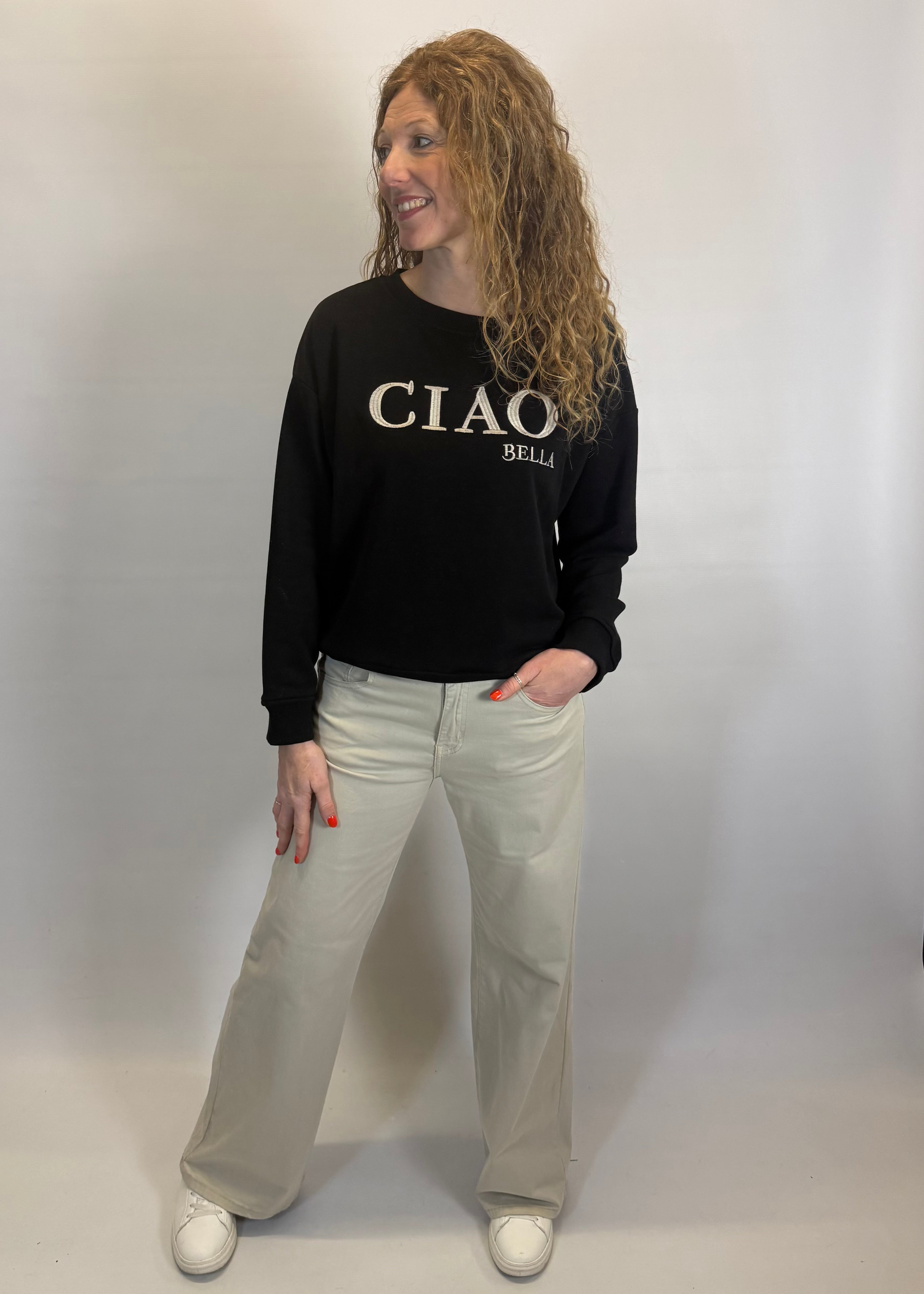 Person wearing a black sweatshirt with 'CIAO BELLI' text and light-colored pants on a plain background