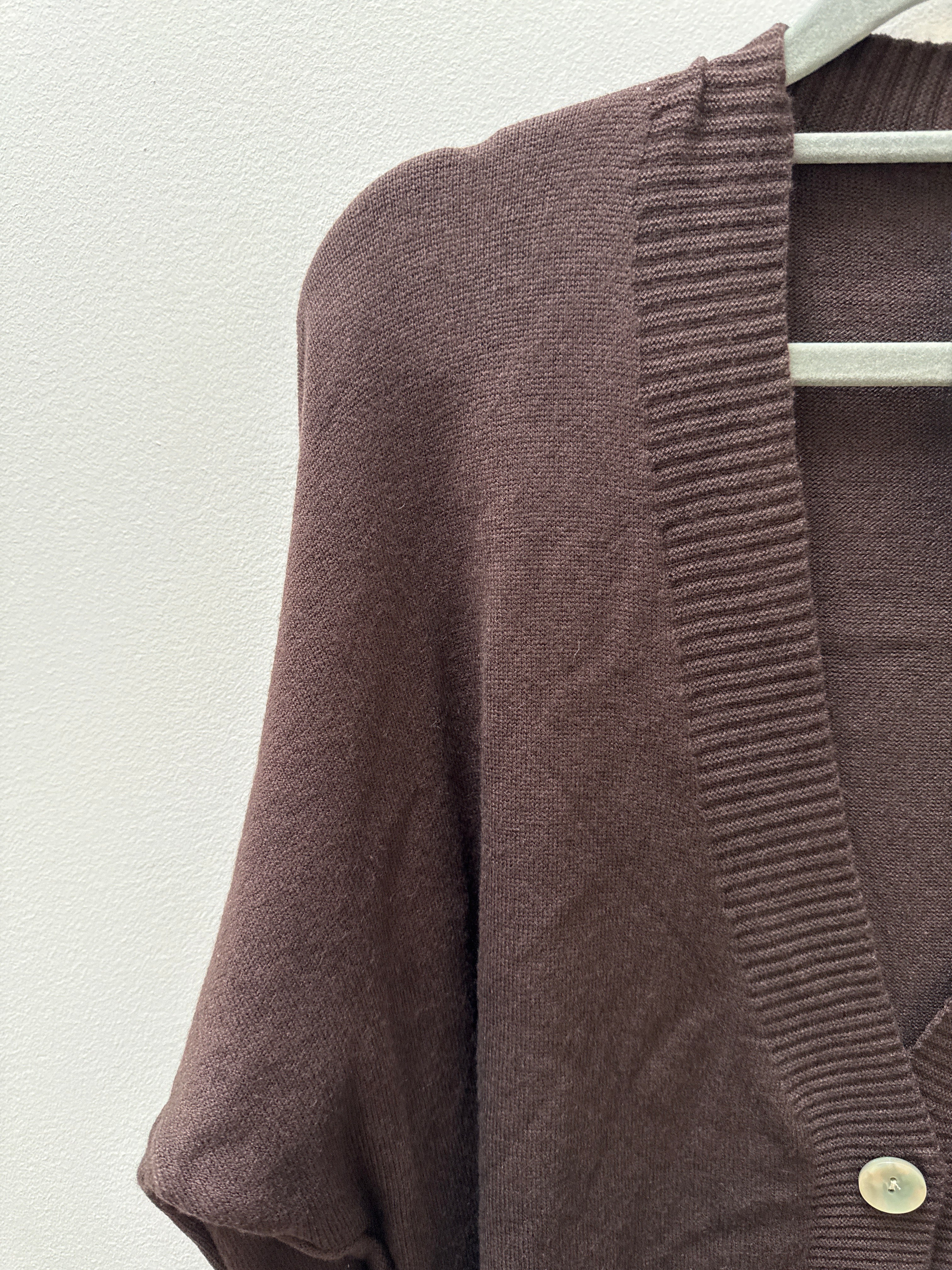 A close-up of a brown knit cardigan sweater hanging on a white hanger against a textured off-white wall. The cardigan features a ribbed V-neck trim and a single visible light-colored button.