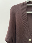 A close-up of a brown knit cardigan sweater hanging on a white hanger against a textured off-white wall. The cardigan features a ribbed V-neck trim and a single visible light-colored button.