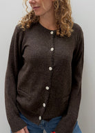 A woman wearing a dark brown knit cardigan sweater with decorative white buttons, paired with blue jeans, shown against a plain light gray wall.