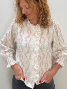 A woman with curly light brown hair wearing a cream-colored long-sleeve blouse with a pink floral pattern and dark gray pants, standing against a plain light gray wall.