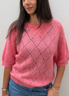 A woman wearing a pink fuzzy knit sweater with a dark gray diamond lattice pattern, paired with blue jeans, standing against a plain light gray wall.