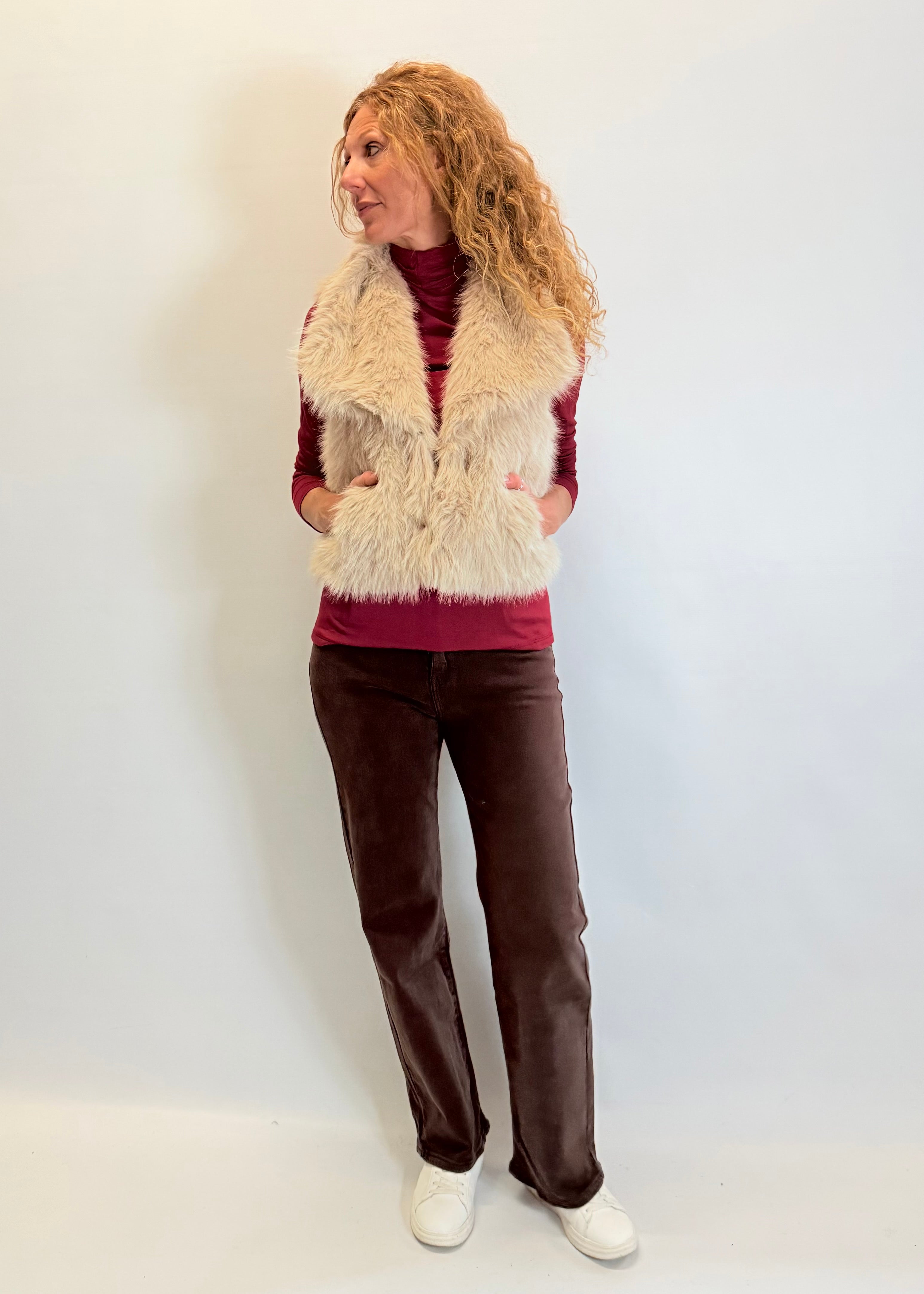 Woman wearing a fur vest, red top, brown pants, and white shoes against a plain background