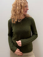 A woman with curly blonde hair wearing an olive green cable-knit sweater and beige pants, standing against a plain light gray wall.