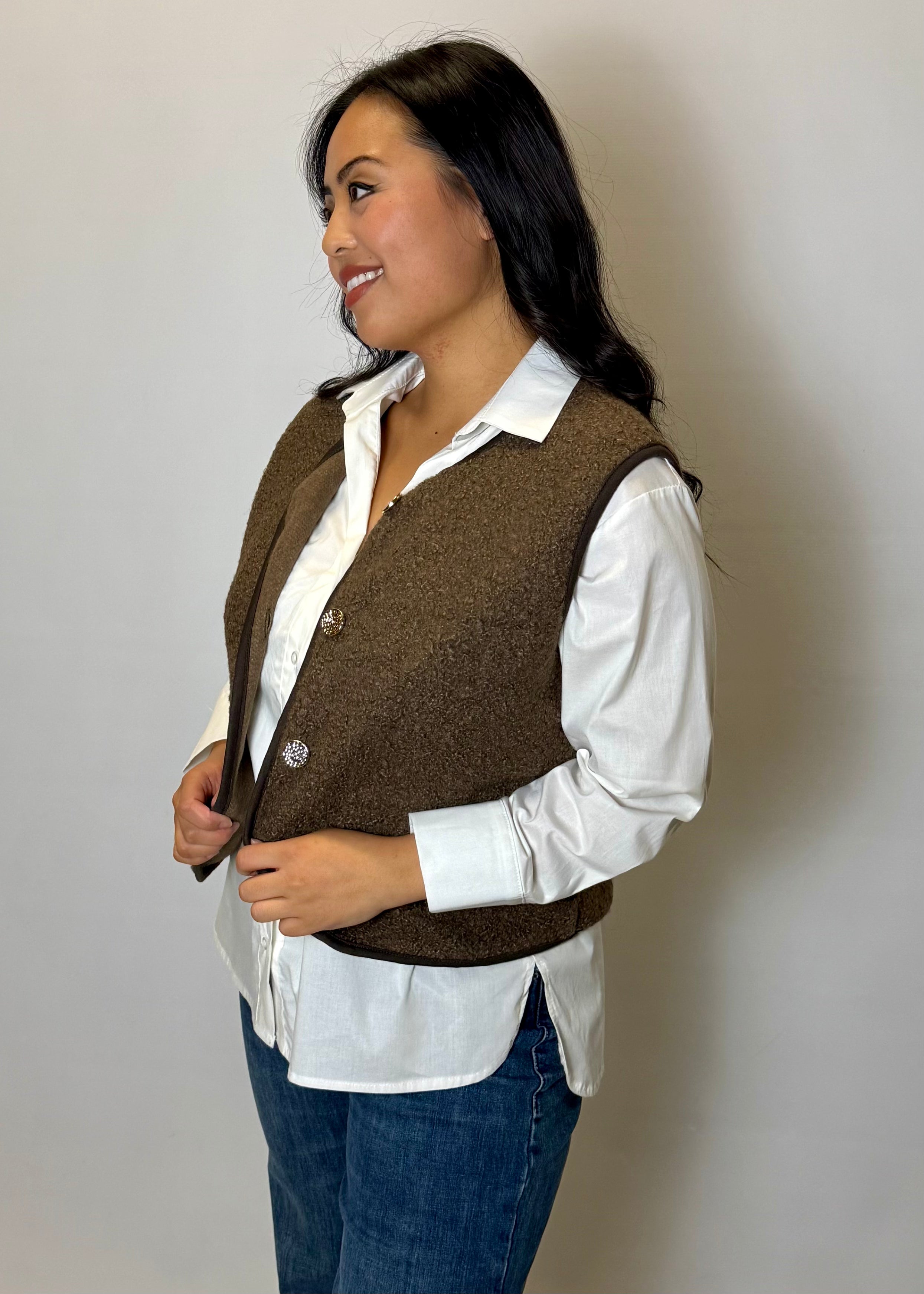 Woman wearing a brown vest over a white shirt and blue jeans against a plain background