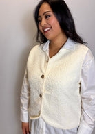Woman wearing a cream-colored vest over a white shirt against a plain background