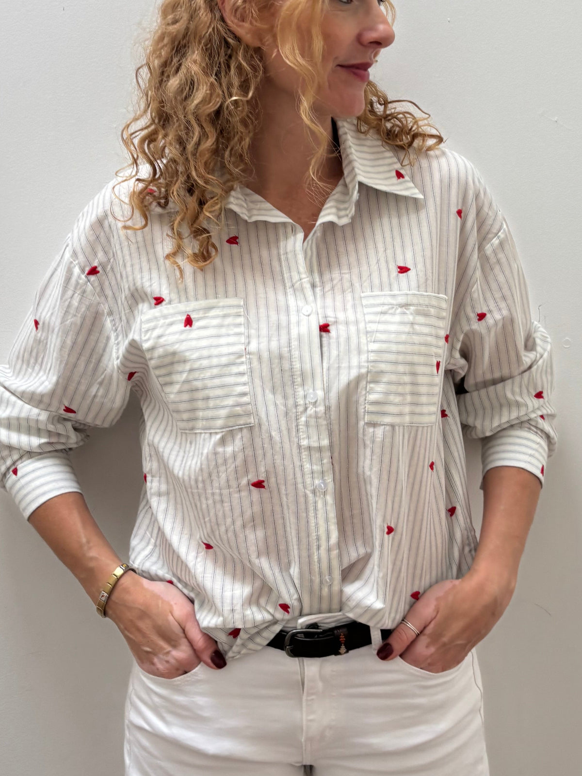 A woman with curly blonde hair stands against a plain light gray wall wearing a white long-sleeve button-up shirt with thin vertical gray stripes and small red embroidered motifs, paired with white pants and a black belt.