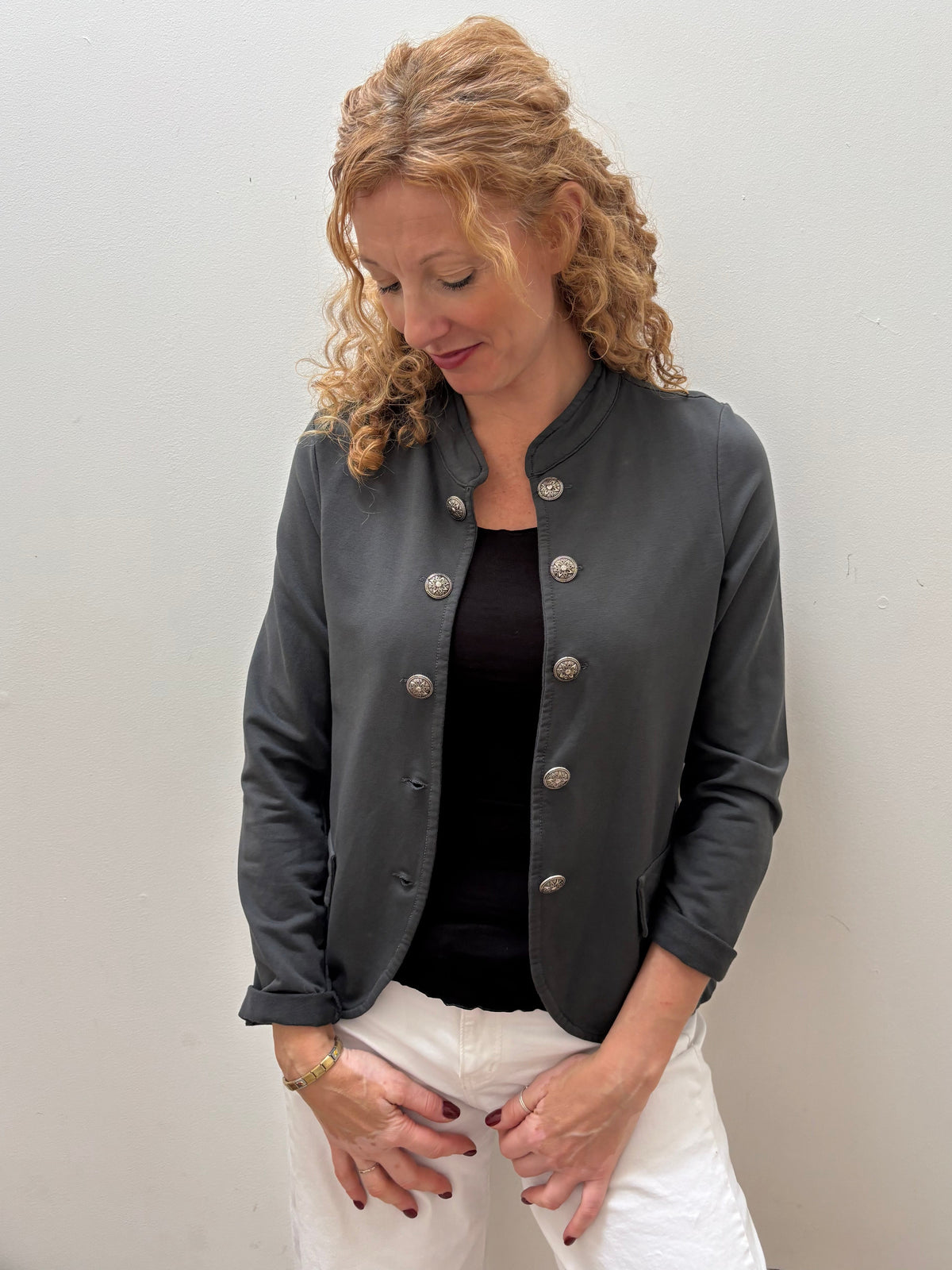 A woman with curly blonde hair wearing a dark gray leather jacket with silver buttons, a black top, and white pants, standing against a plain light gray wall.