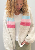 A woman wearing a fuzzy cream-colored cardigan with horizontal pastel stripes in light blue, pink, and magenta, layered over a white top and olive green pants.