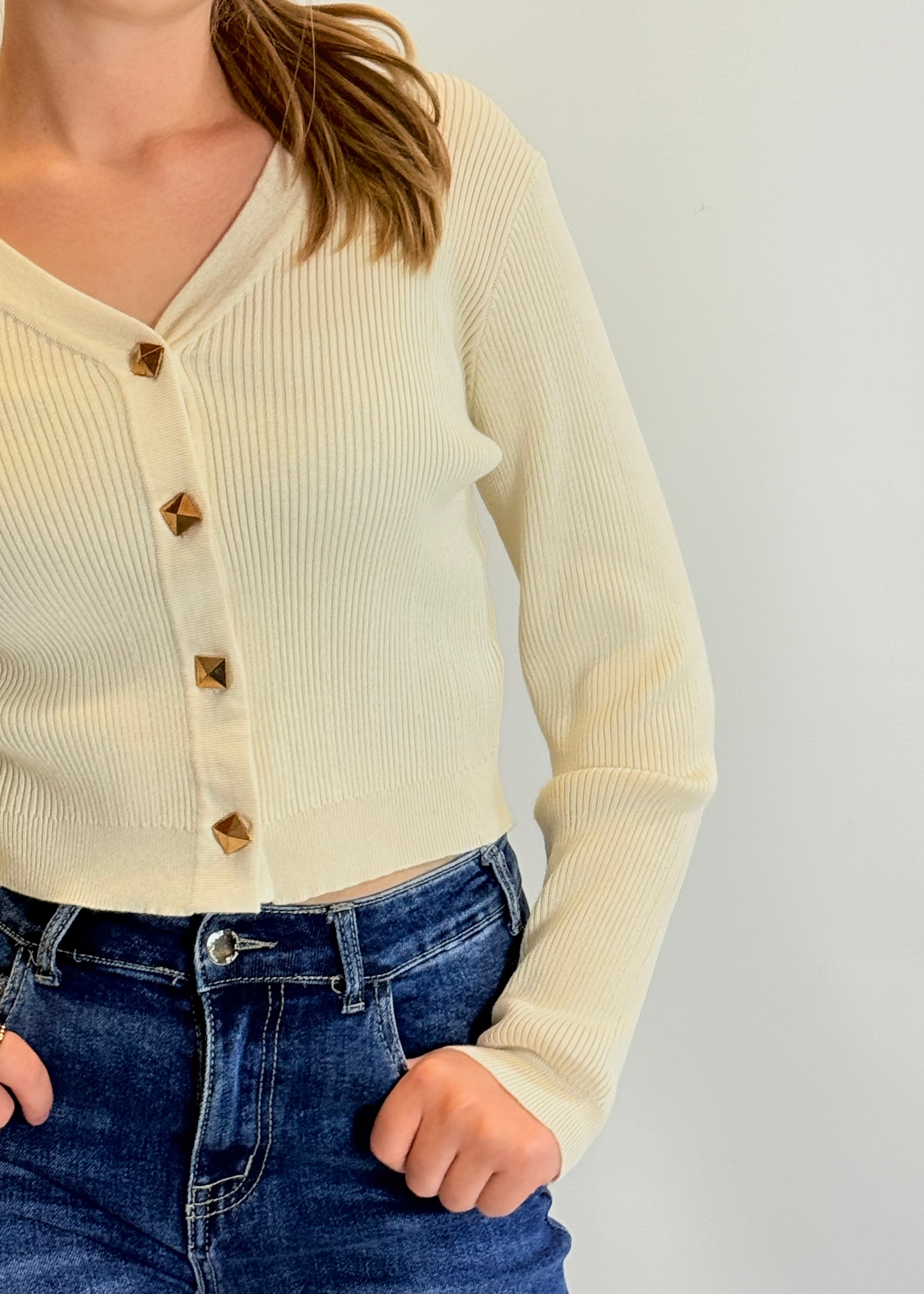 Person wearing a cream ribbed cardigan with gold buttons and blue jeans on a light gray background