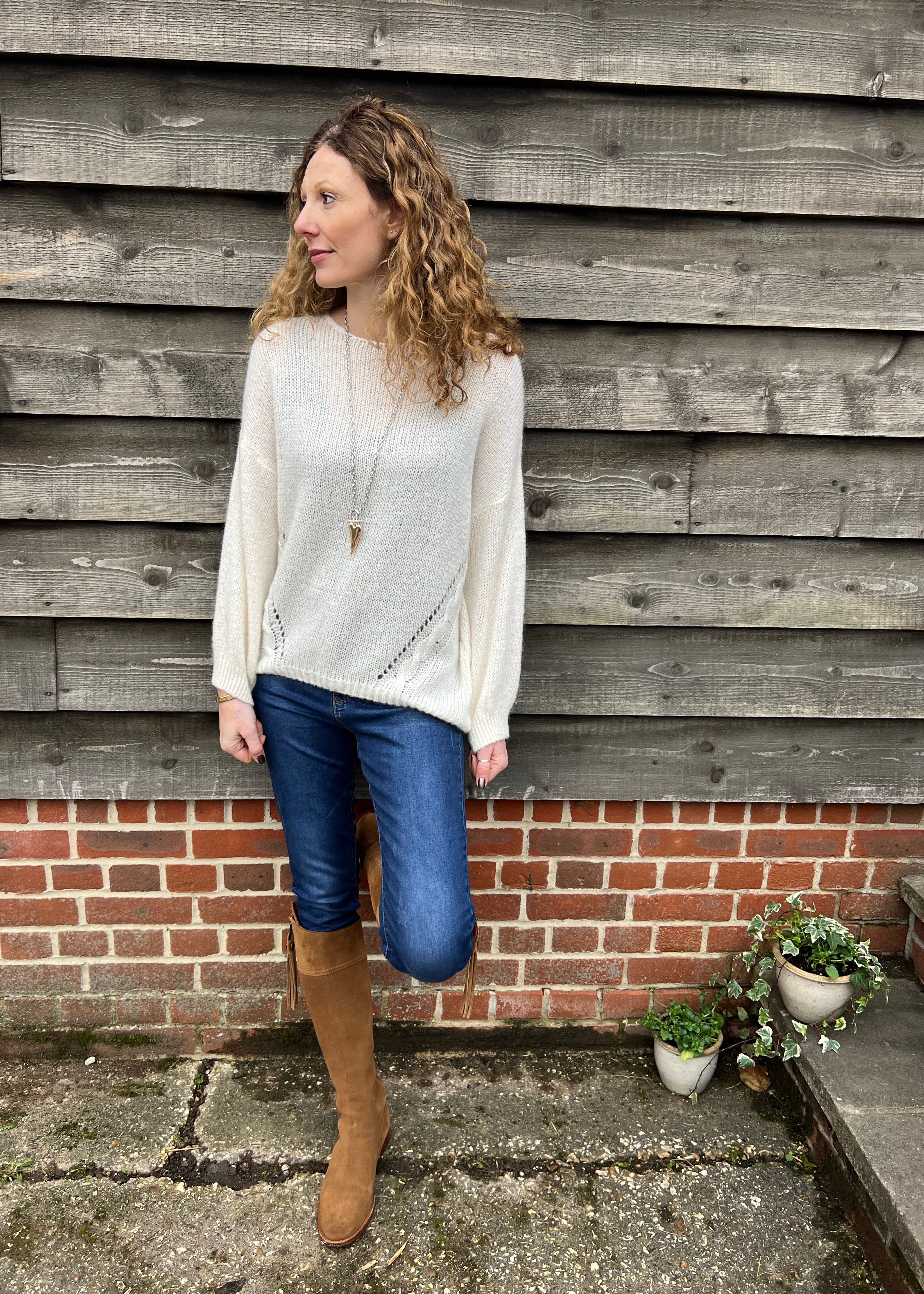 A woman standing outdoors wearing a white knitted jumper with 3/4 length sleeves and brown boots, with a wooden background and a potted plant beside her.