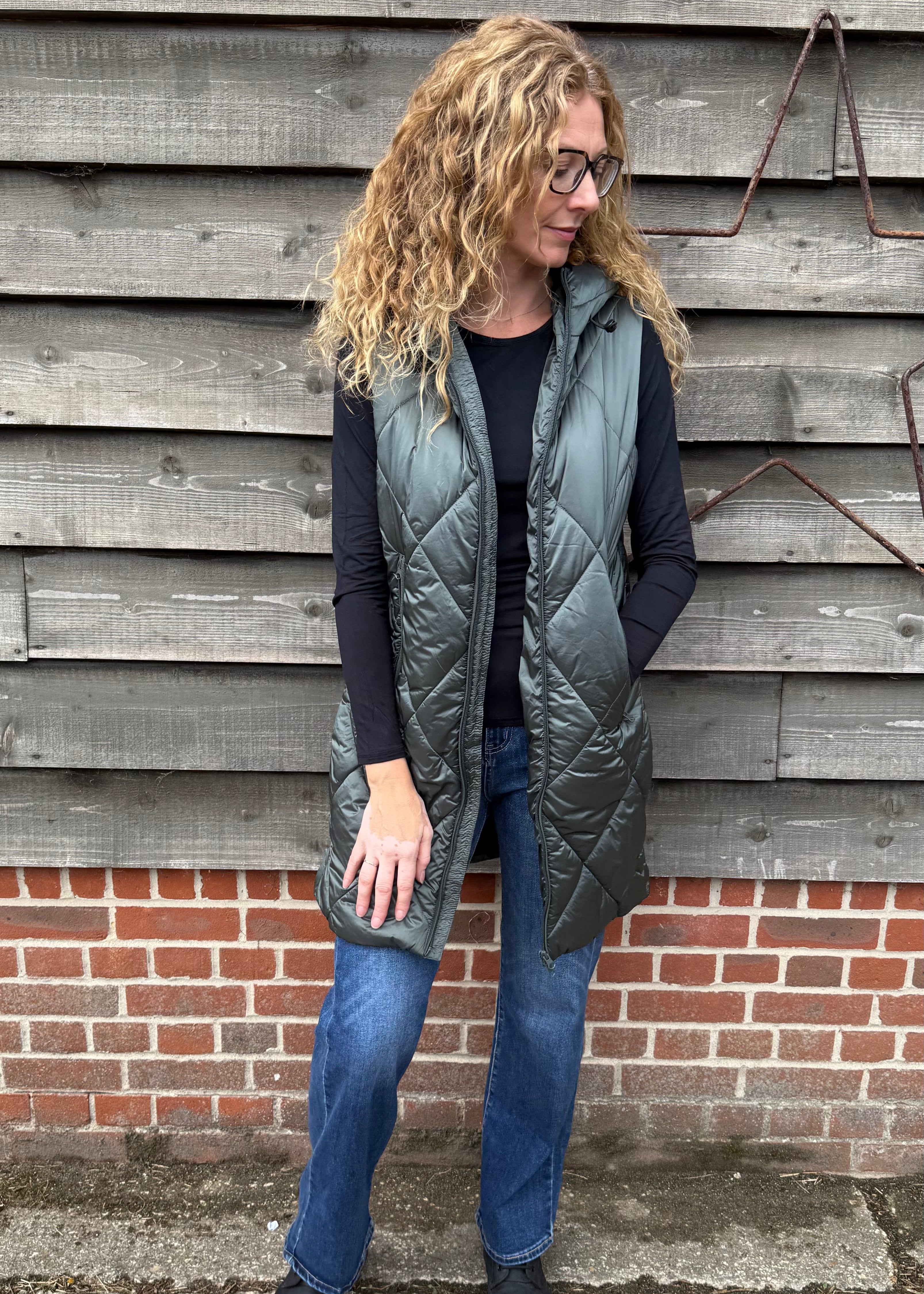 Woman wearing a long gray quilted vest standing against a wooden wall.