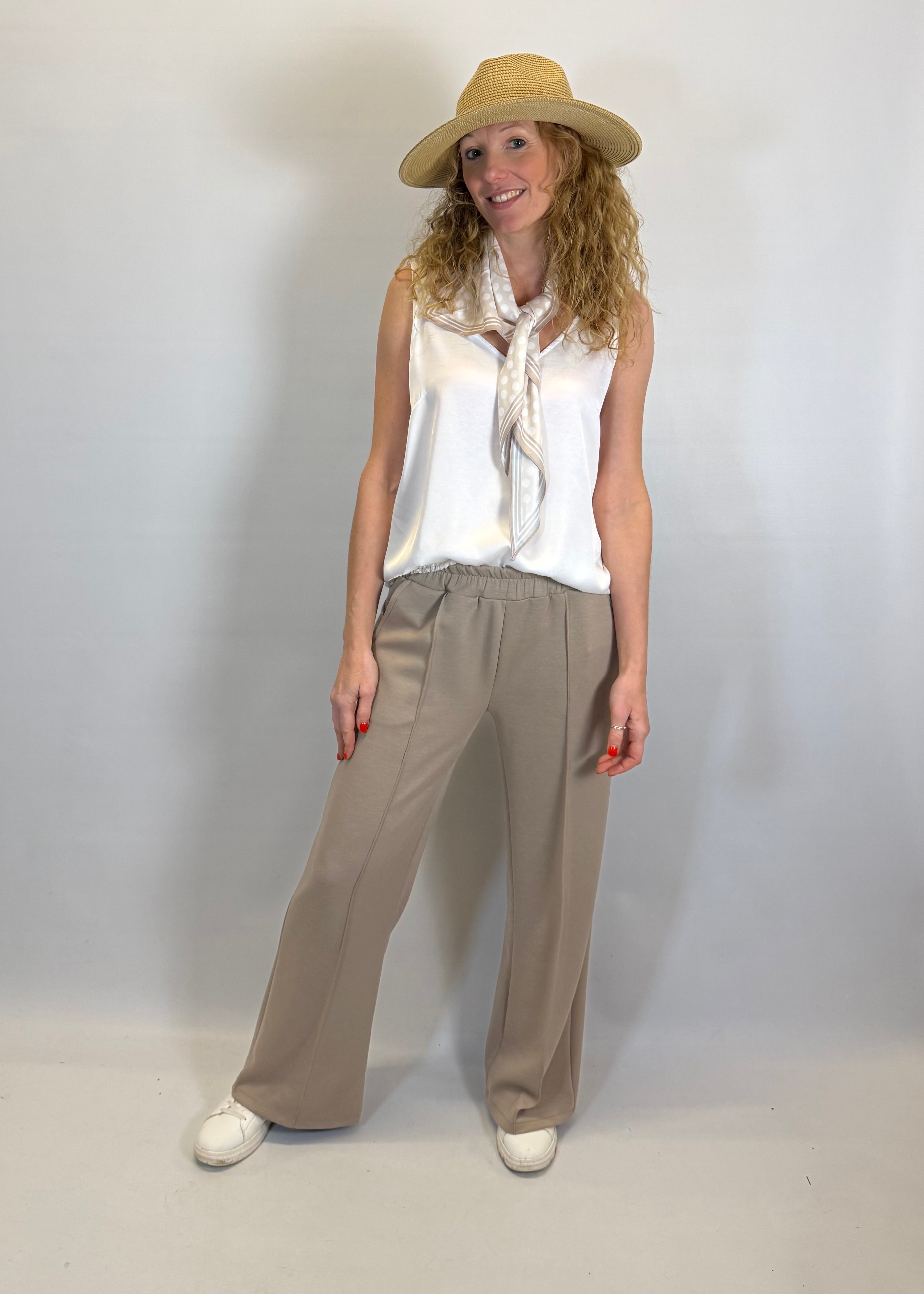 Woman wearing a white sleeveless top, beige wide-leg pants, and a beige hat against a plain background