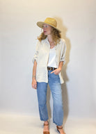 Person wearing a beige hat, light-colored shirt, white top, blue jeans, and brown sandals against a white background