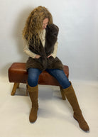 Woman sitting on a brown bench wearing a fur vest and knee-high boots against a white background