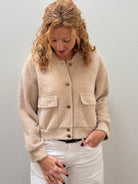 A woman with blonde curly hair wearing a beige fuzzy cropped cardigan with button details and white pants, standing against a plain light gray wall.