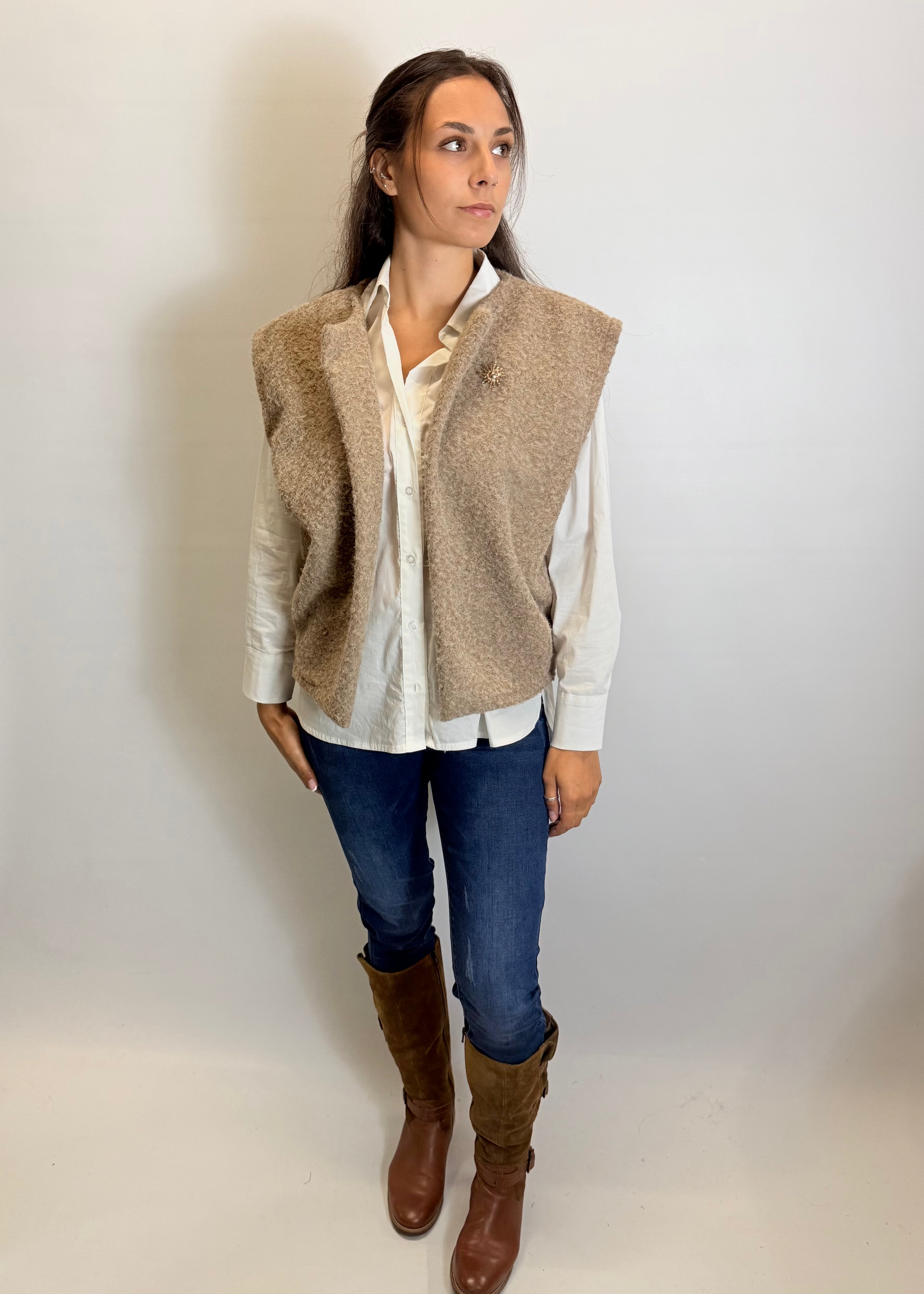 Woman wearing a beige vest over a white shirt, blue jeans, and brown boots on a plain background