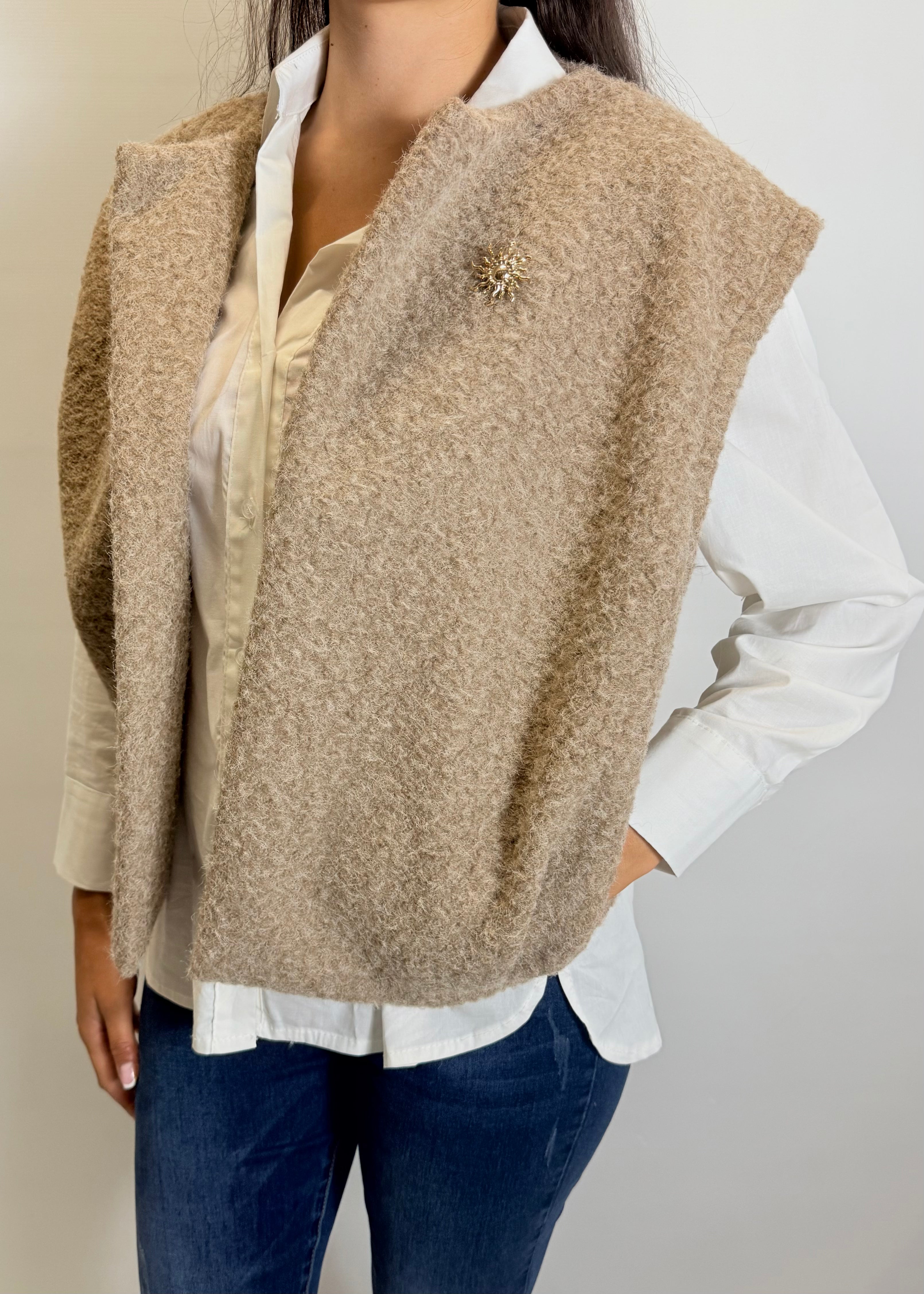 Person wearing a beige vest over a white shirt and blue jeans on a plain background