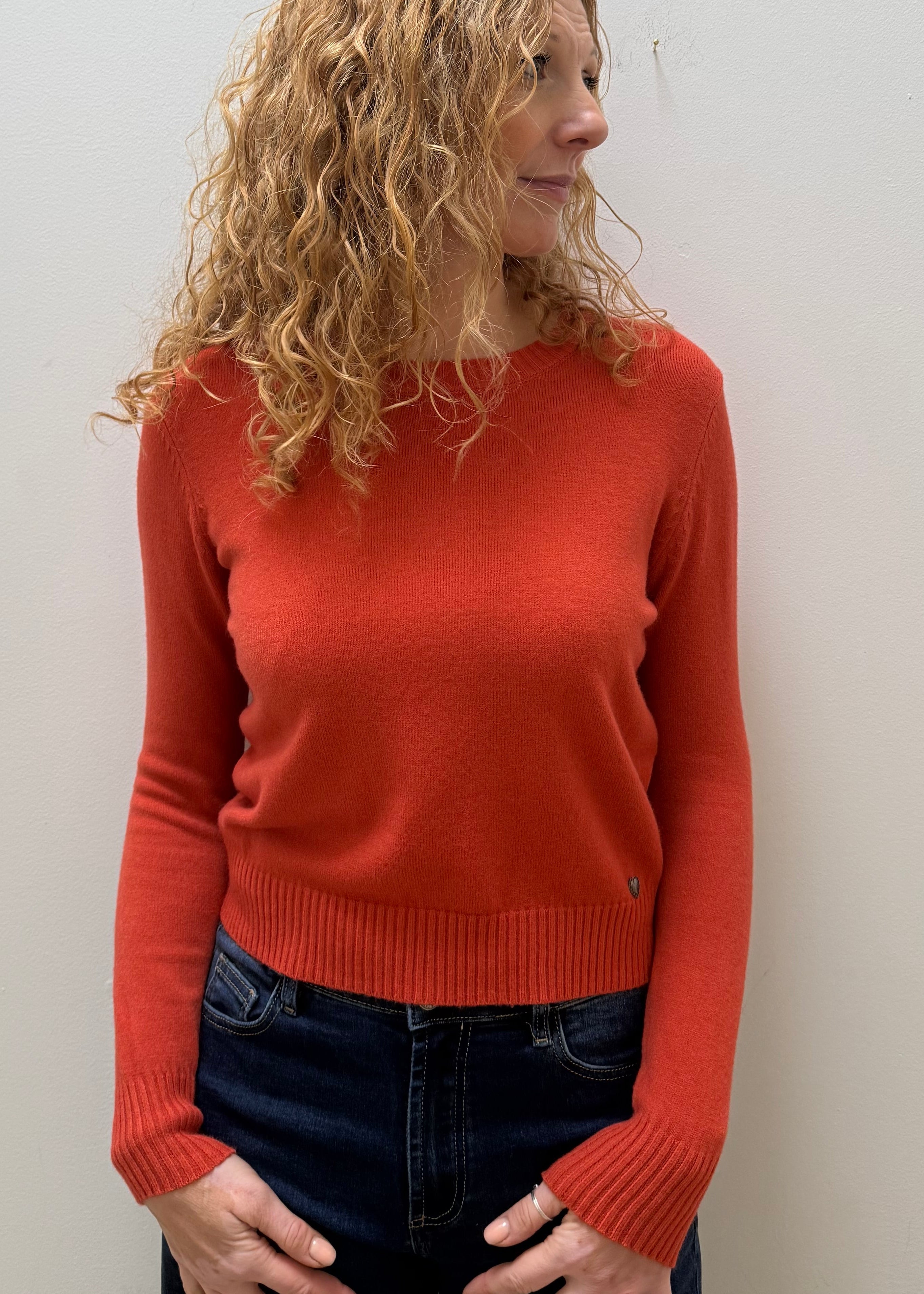 A woman with curly blonde hair wearing a rust-orange long-sleeve knit sweater and dark blue jeans, standing against a plain off-white wall.