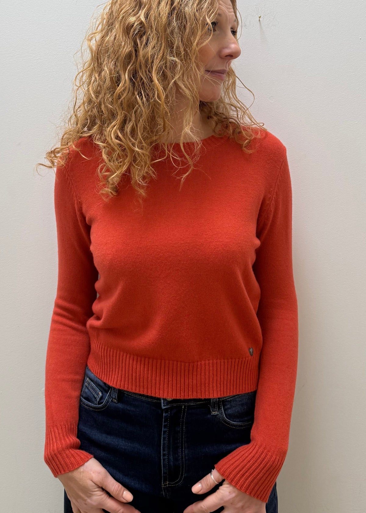 A woman with curly blonde hair wearing a rust-orange long-sleeve knit sweater and dark blue jeans, standing against a plain off-white wall.
