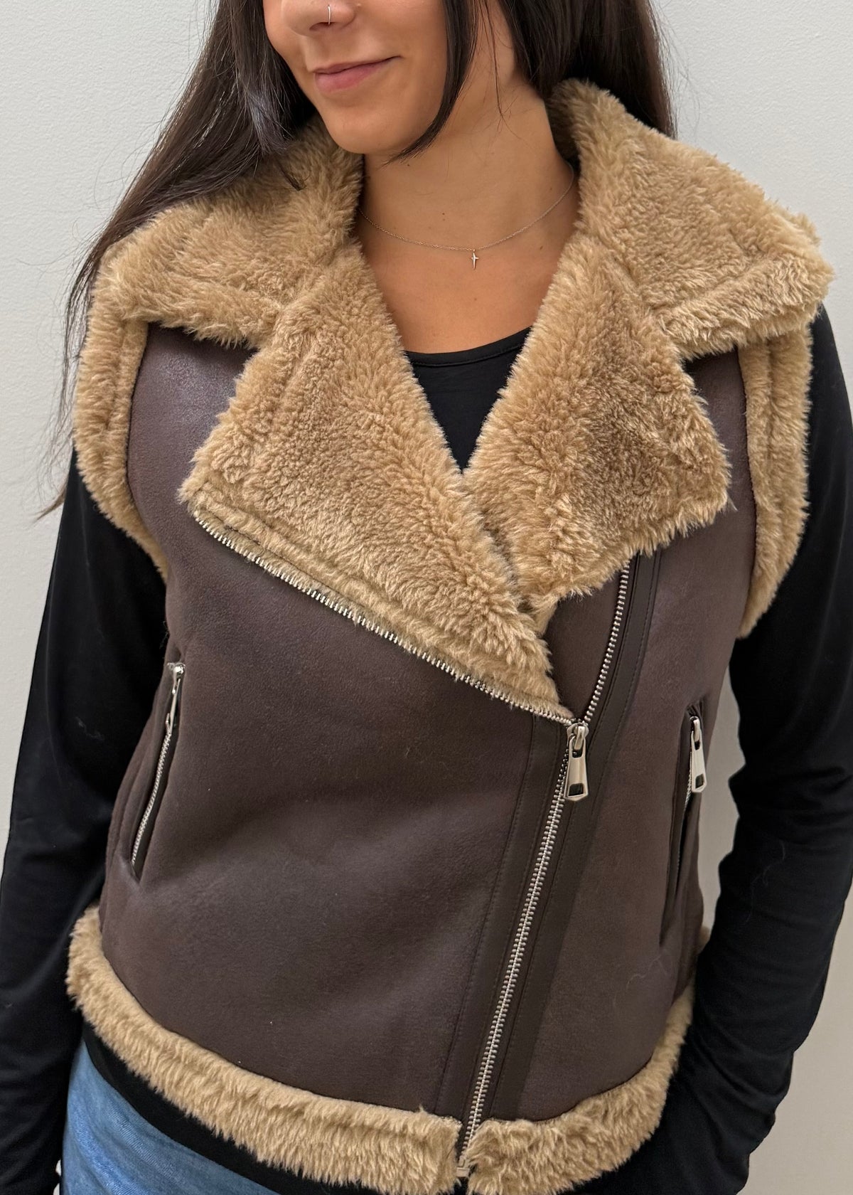 A close-up of a person wearing a brown faux-leather aviator-style vest with a thick beige sherpa collar and lining, over a black long-sleeve top.