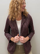 A woman with curly blonde hair wearing a textured plum purple blazer over a white top and khaki pants, standing against a plain light gray wall.