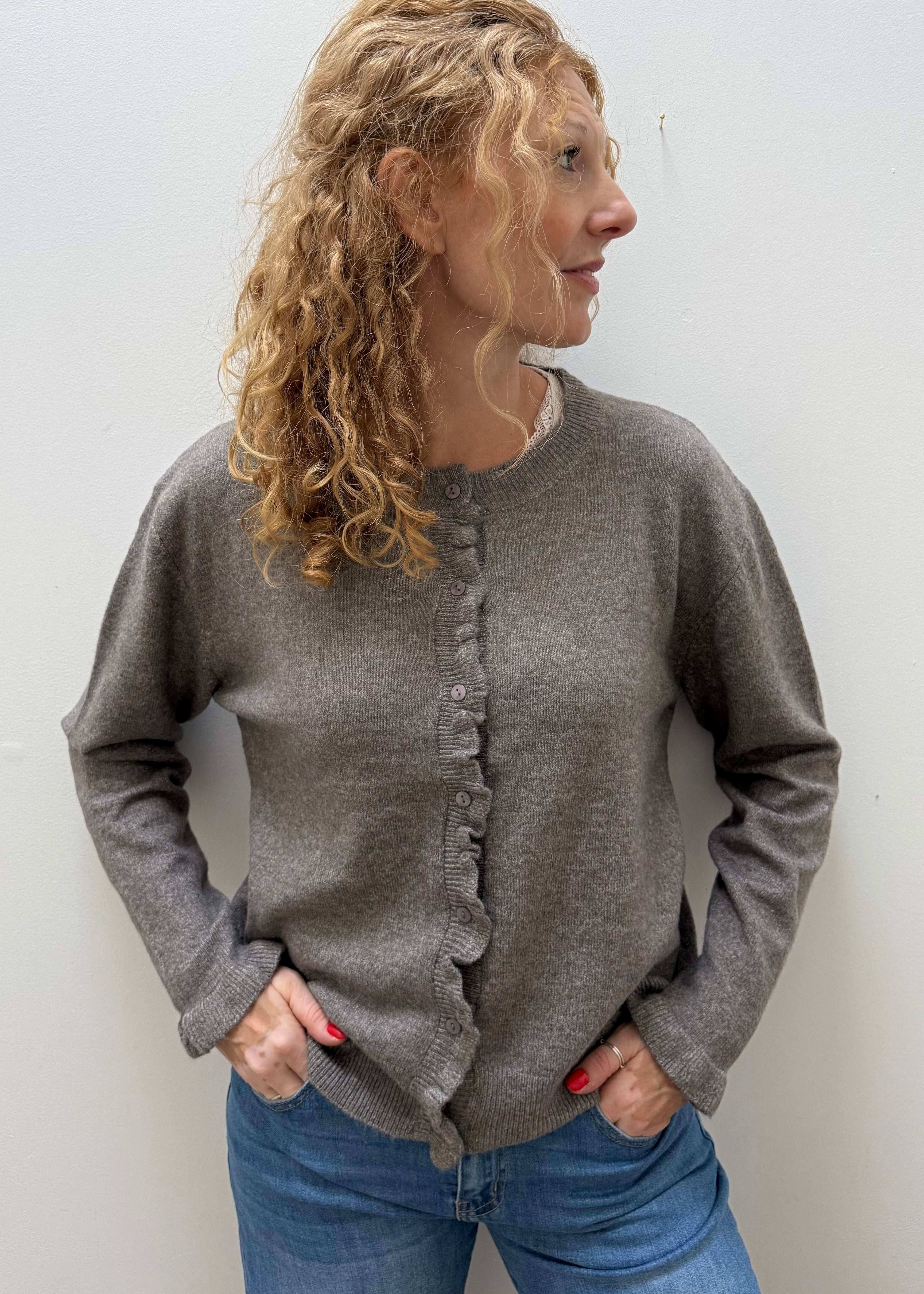 A woman with curly blonde hair wearing a gray knit cardigan sweater with ruffle trim down the front and blue jeans, standing against a plain light gray wall.