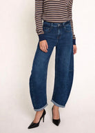 A model wearing dark blue wide-leg cuffed denim jeans, paired with a brown and beige striped long-sleeve top and black pointed-toe heels, standing against a plain off-white background.