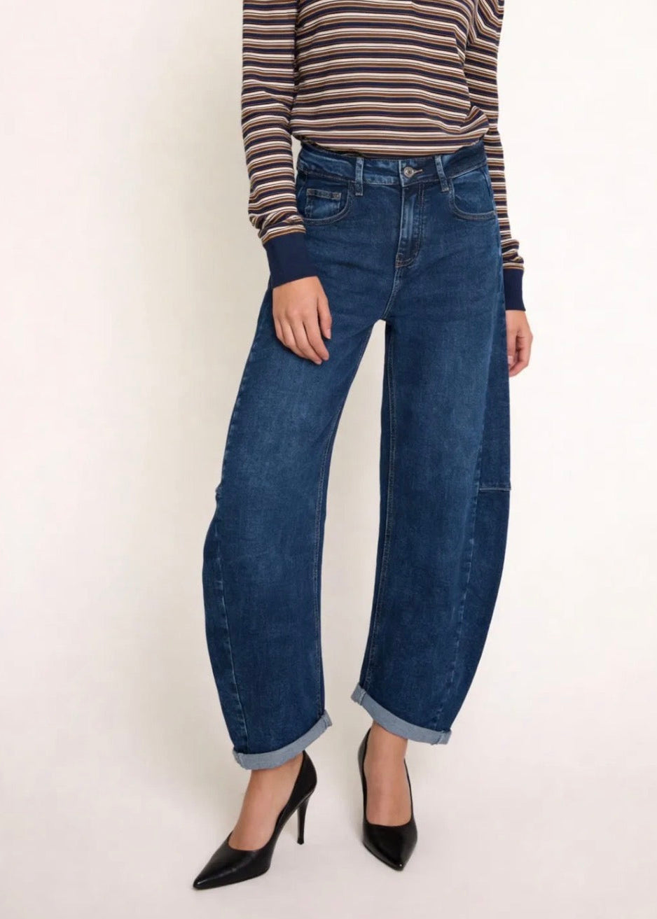 A model wearing dark blue wide-leg cuffed denim jeans, paired with a brown and beige striped long-sleeve top and black pointed-toe heels, standing against a plain off-white background.