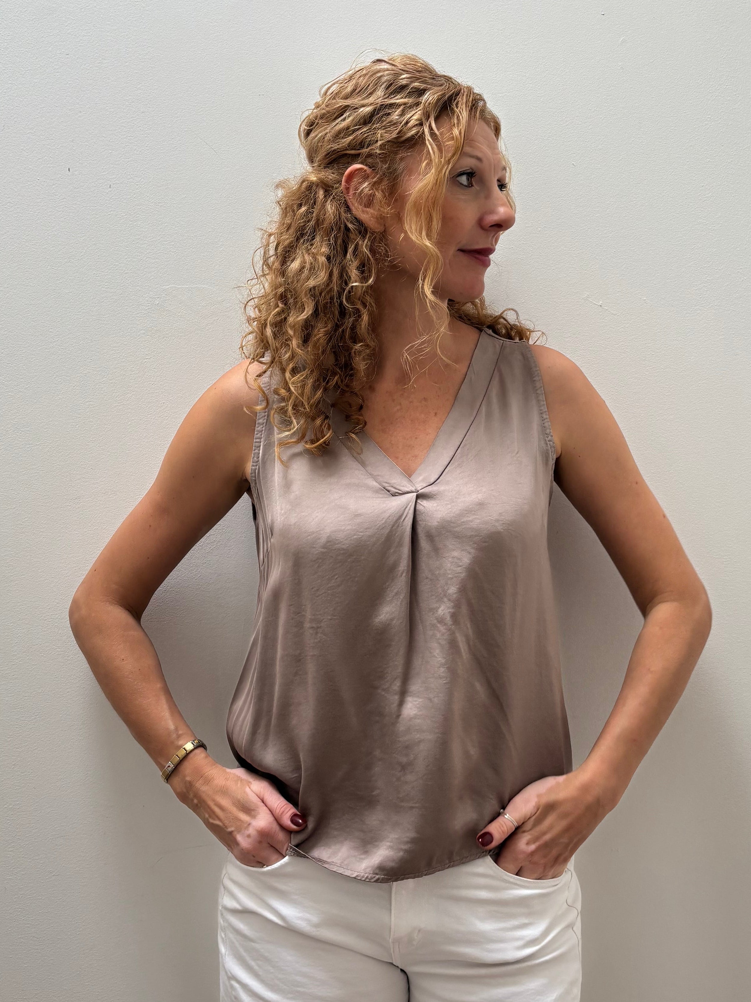 A woman with curly blonde hair stands against a plain light gray wall wearing a sleeveless taupe satin-like top and white pants.