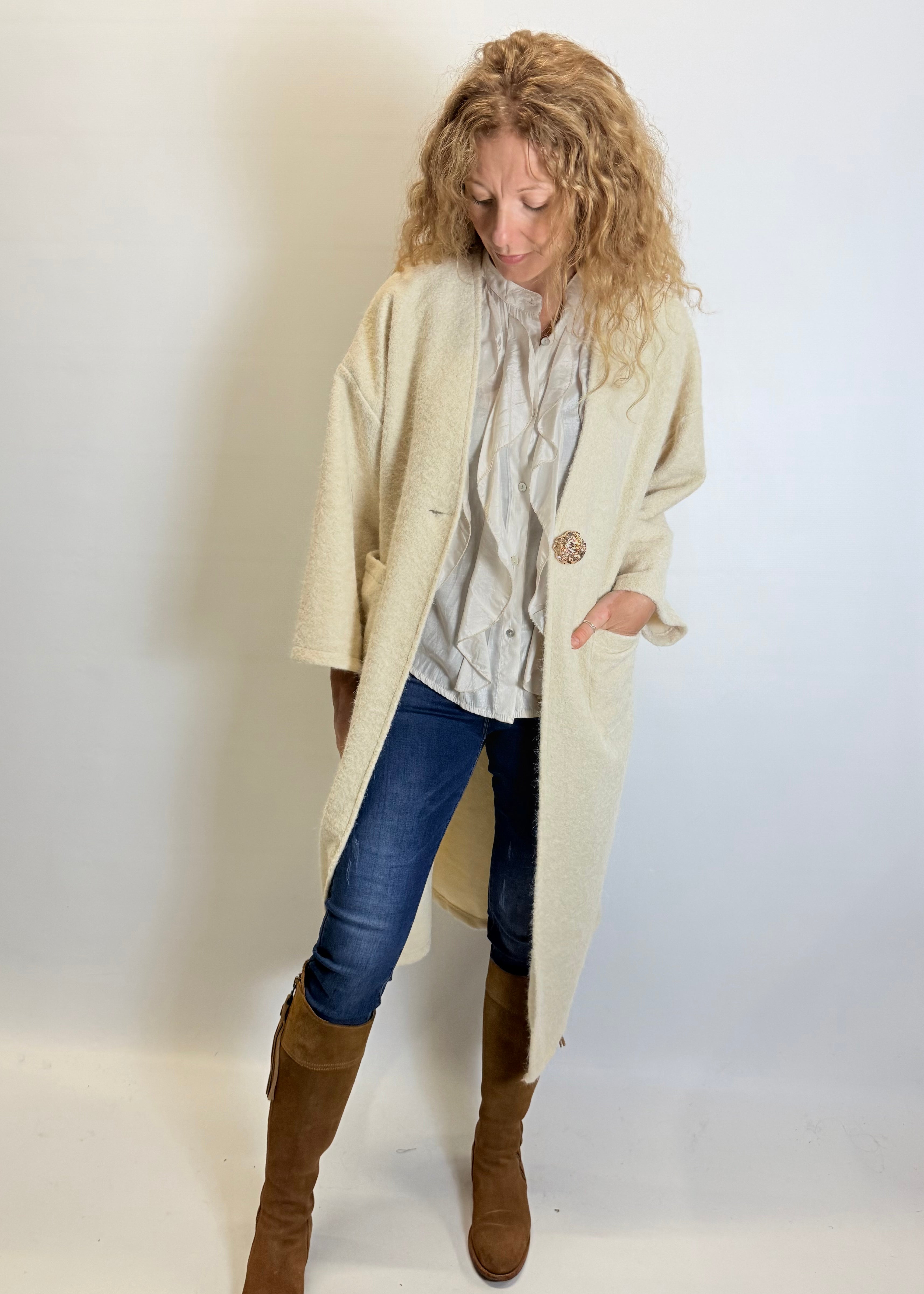 Woman wearing a beige coat, blue jeans, and brown boots against a plain background