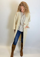 Woman wearing a beige coat, blue jeans, and brown boots against a plain background