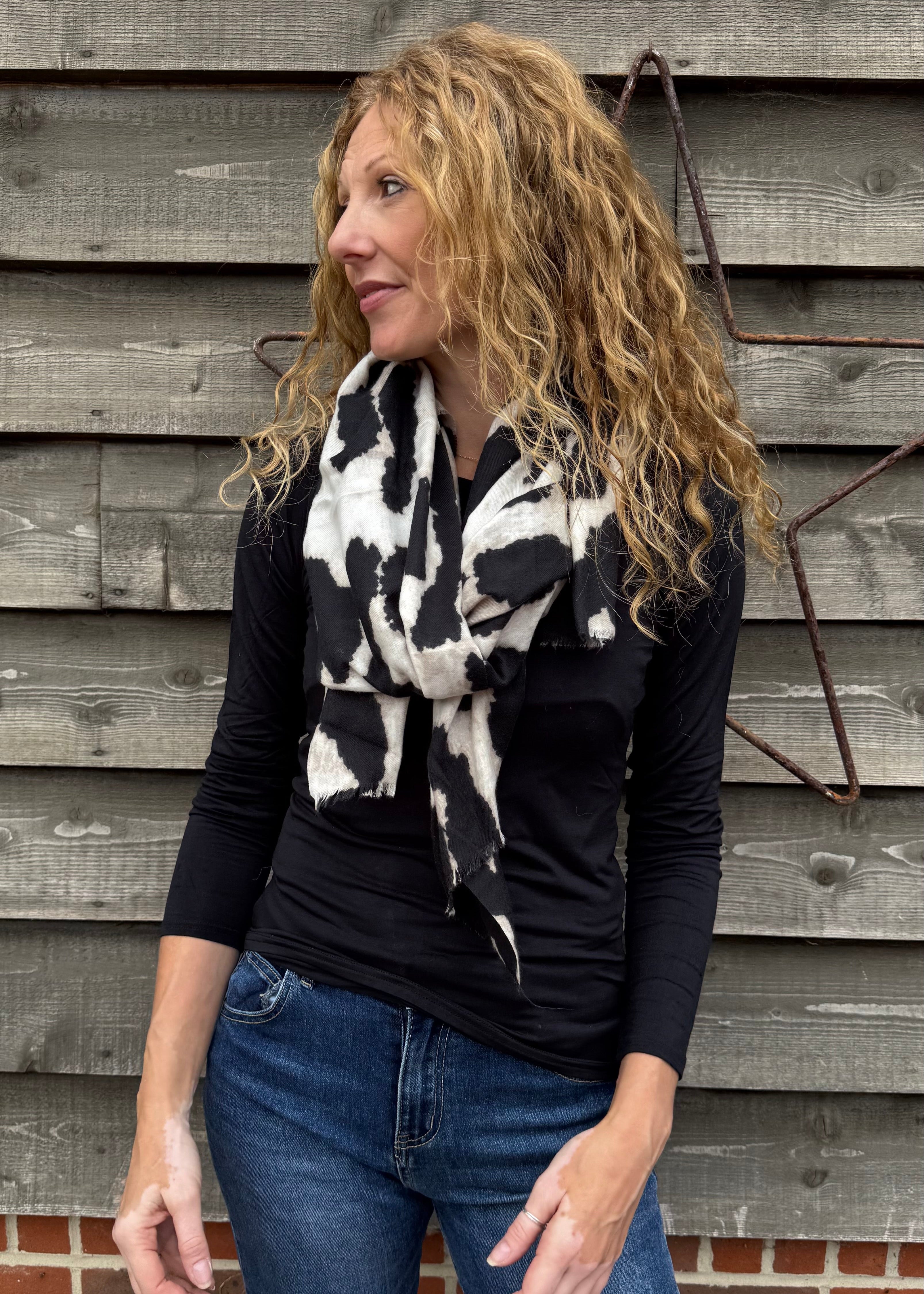 Woman wearing a black top and blue jeans with a black and white patterned scarf against a wooden wall.