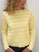 A woman with curly blonde hair wearing a pale yellow knit sweater and dark gray jeans, standing against a plain off-white wall.