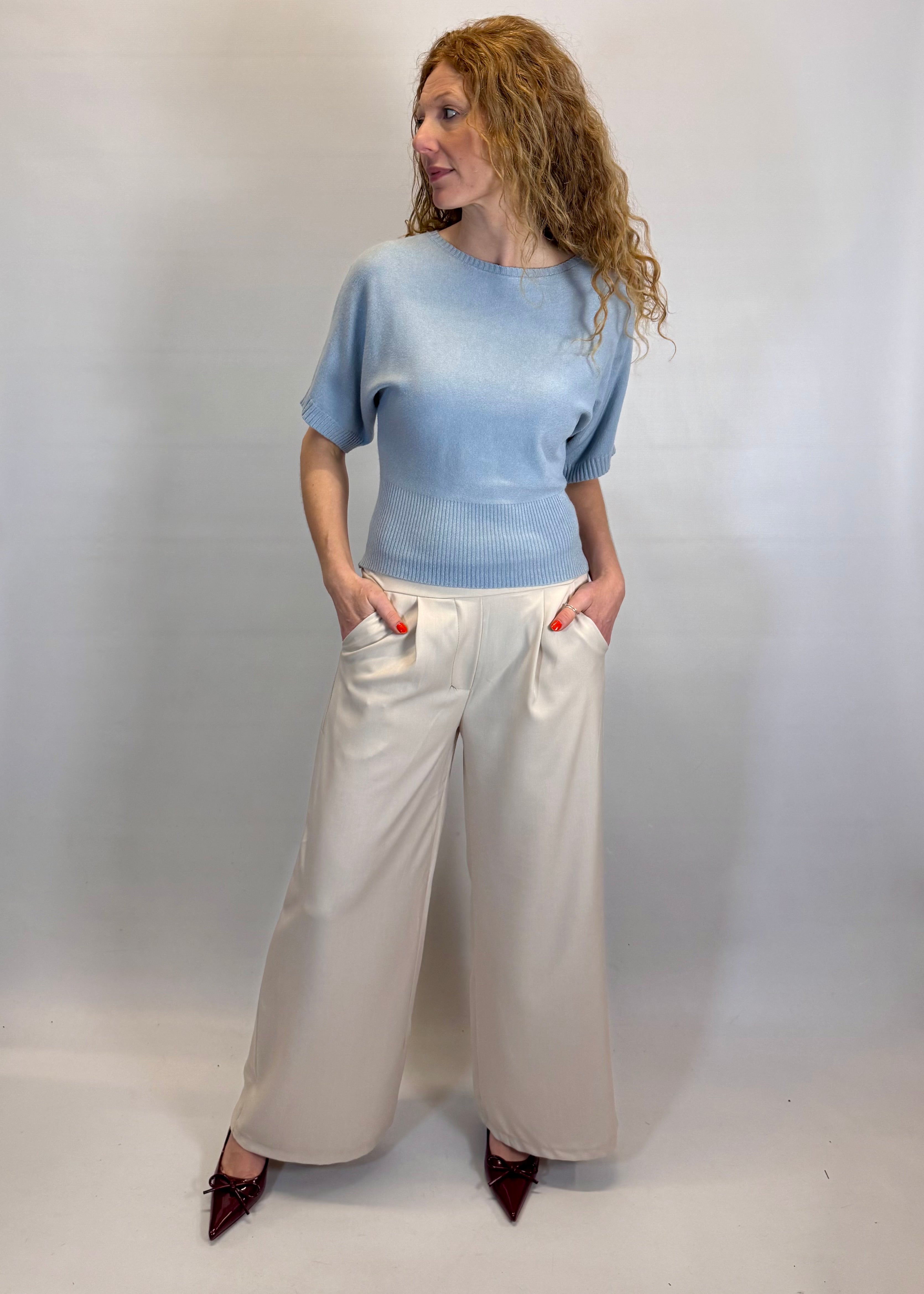 Woman wearing a light blue sweater and white pants against a plain background