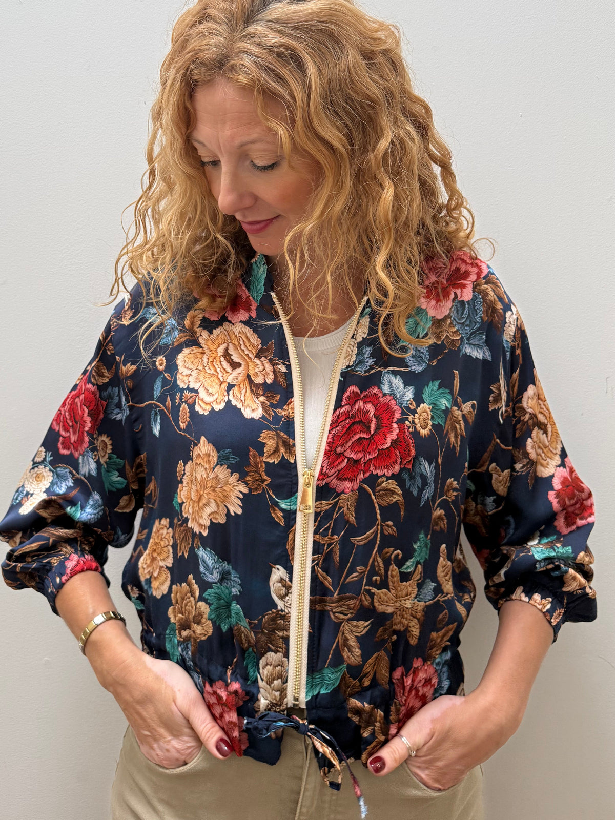 A woman with curly blonde hair wearing a navy blue floral bomber jacket with red, gold, and green flowers, paired with beige pants, standing against a plain light gray wall.
