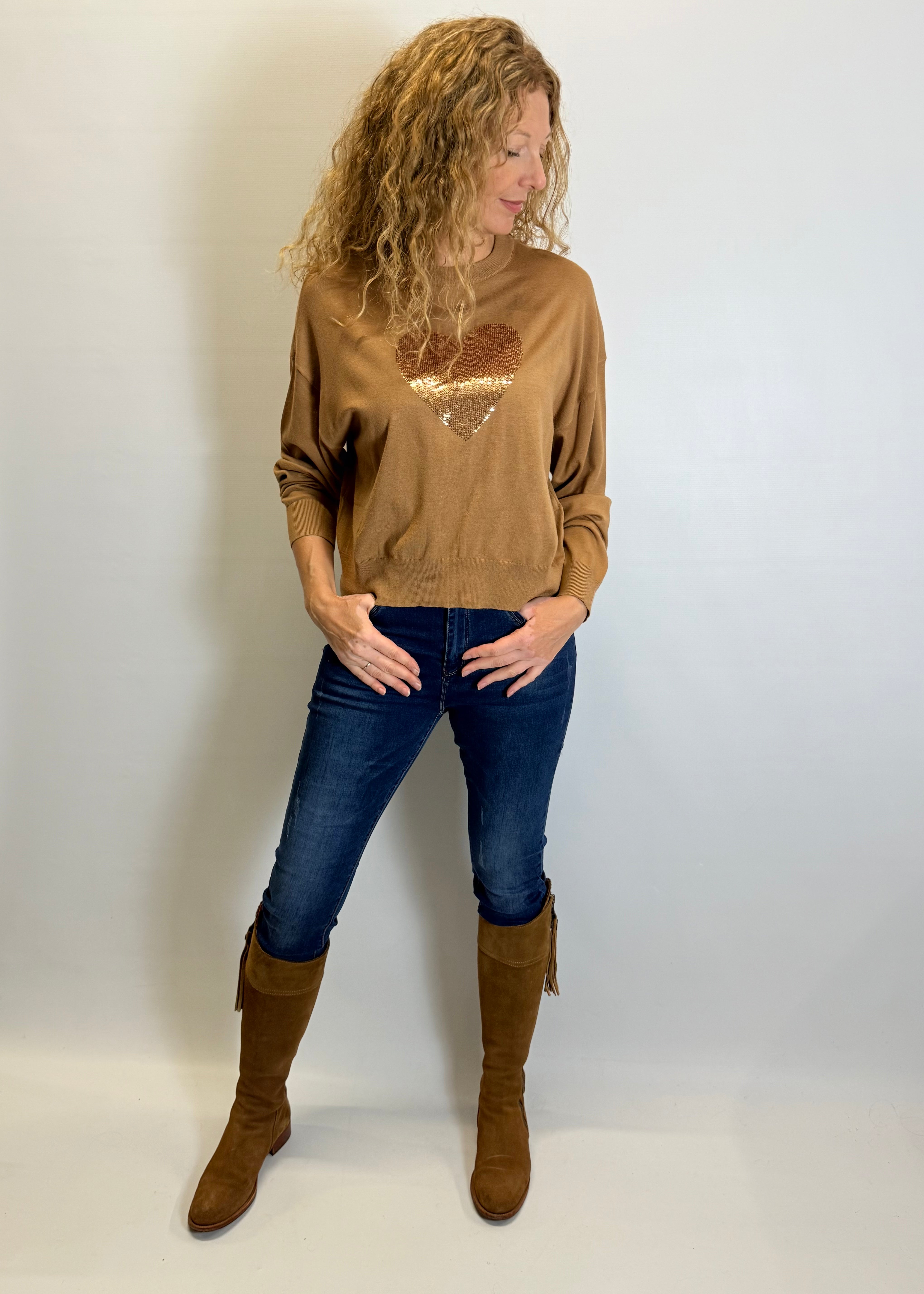 Woman wearing a brown sweater with a heart design, blue jeans, and brown knee-high boots against a white background.