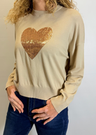 Woman wearing a beige sweater with a gold sequin heart design against a plain background