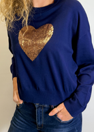 Woman wearing a blue sweater with a gold sequin heart design on a plain background