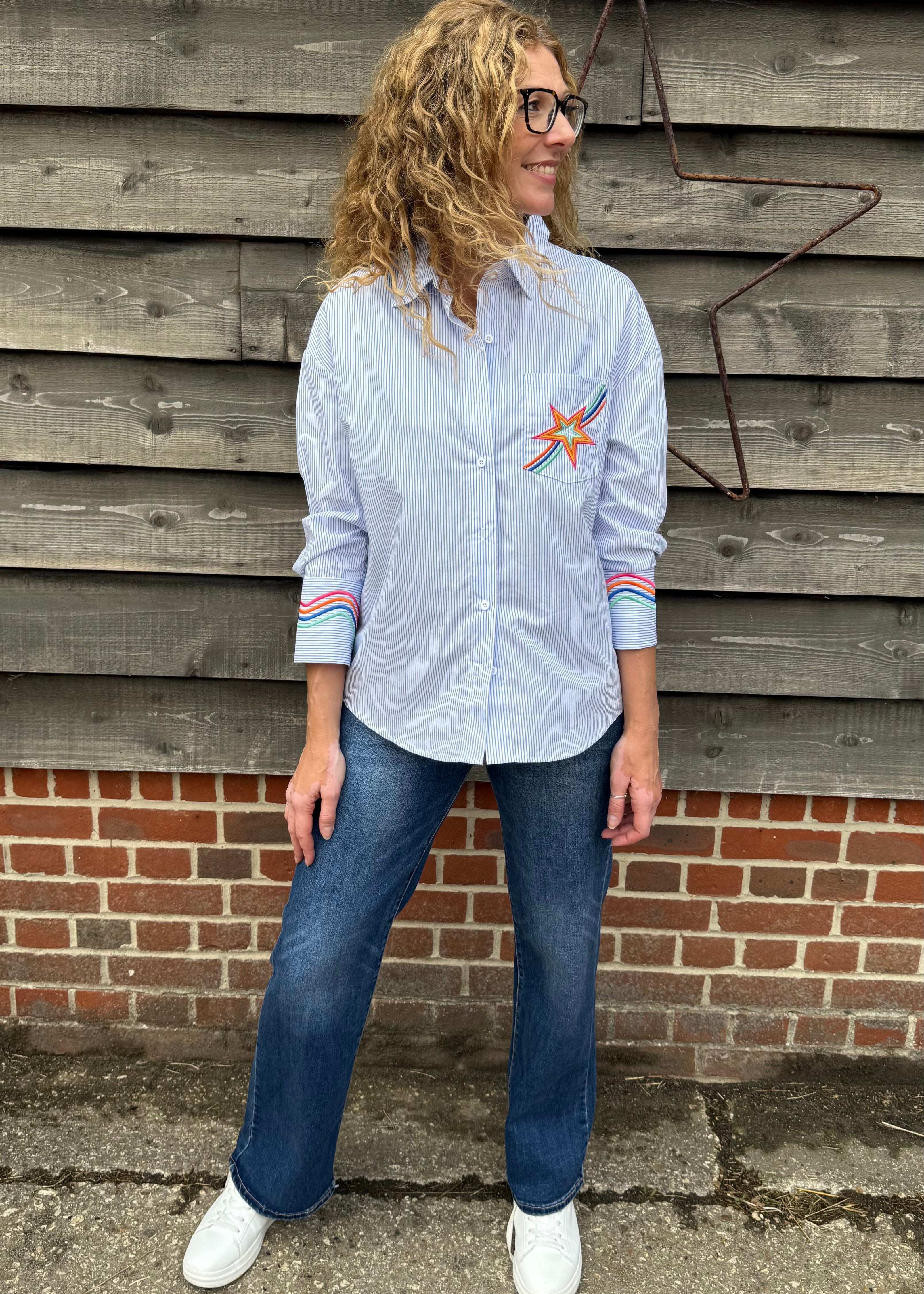 Person wearing a light blue shirt with a star design and jeans, standing against a wooden wall.