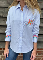 Woman wearing a blue striped shirt with a colorful star design and rainbow sleeves against a wooden wall.