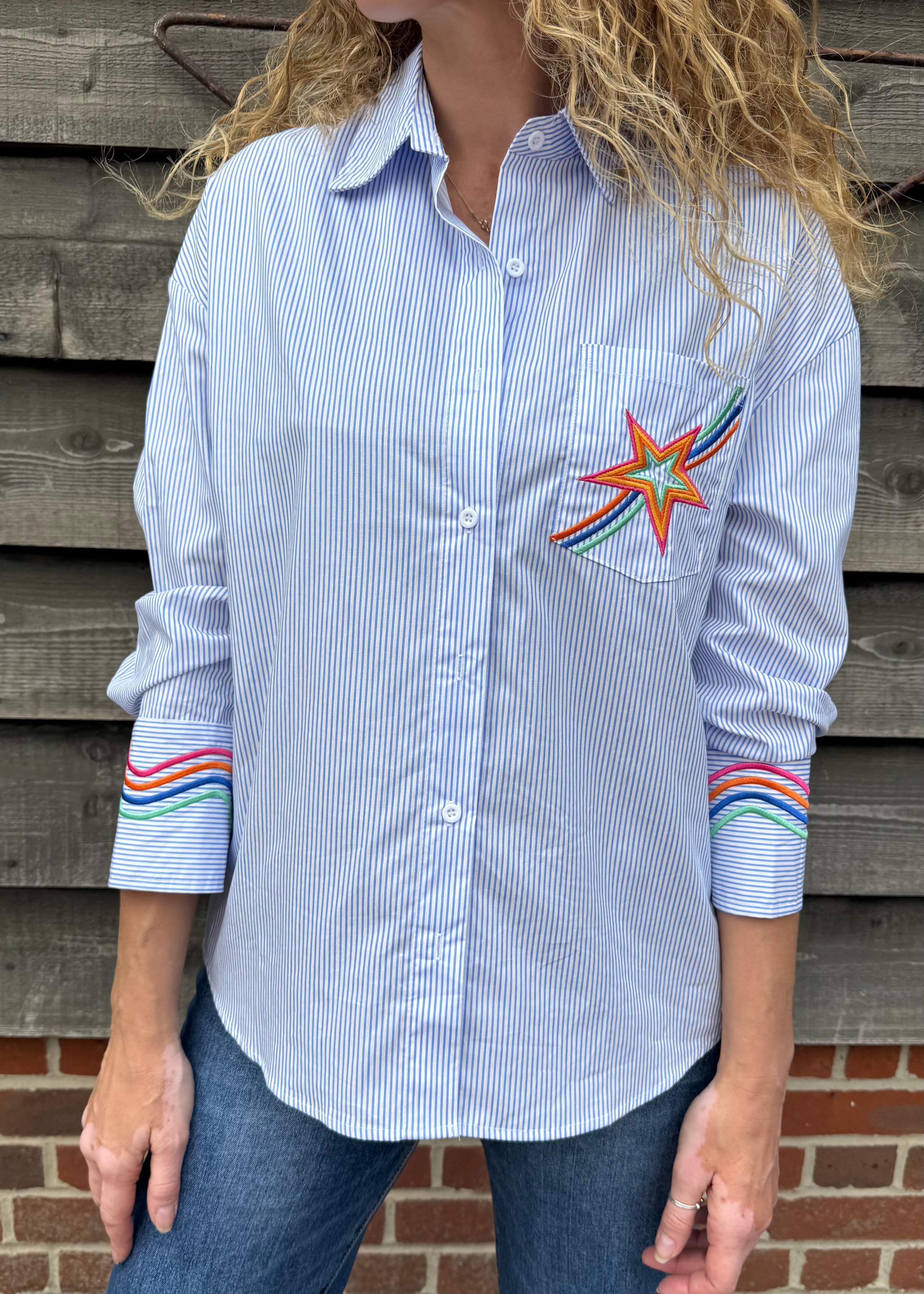 Woman wearing a blue striped shirt with a colorful star design and rainbow sleeves against a wooden wall.