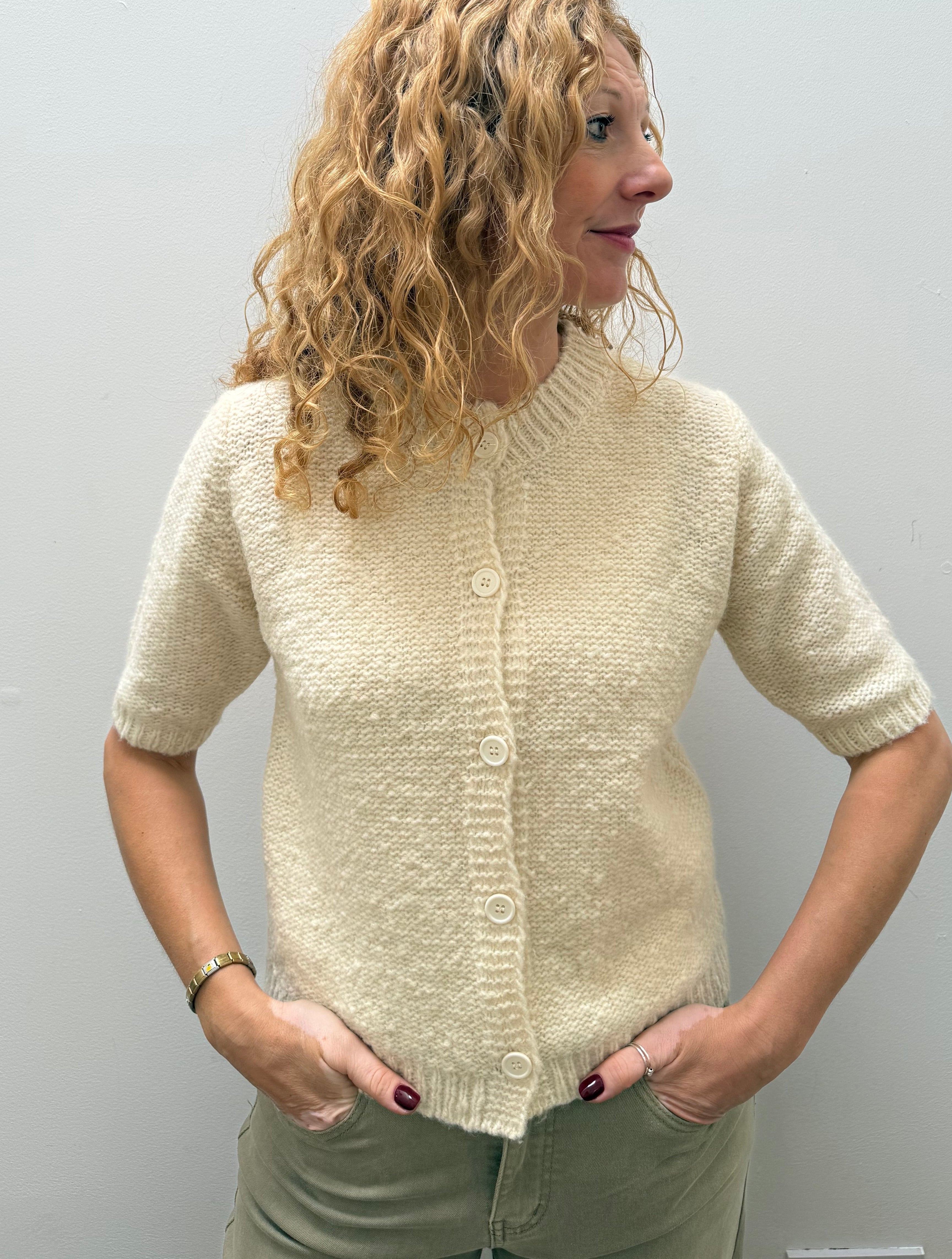 A woman with curly blonde hair wearing a cream-colored short-sleeve knit cardigan and olive green pants, standing against a plain light gray wall.