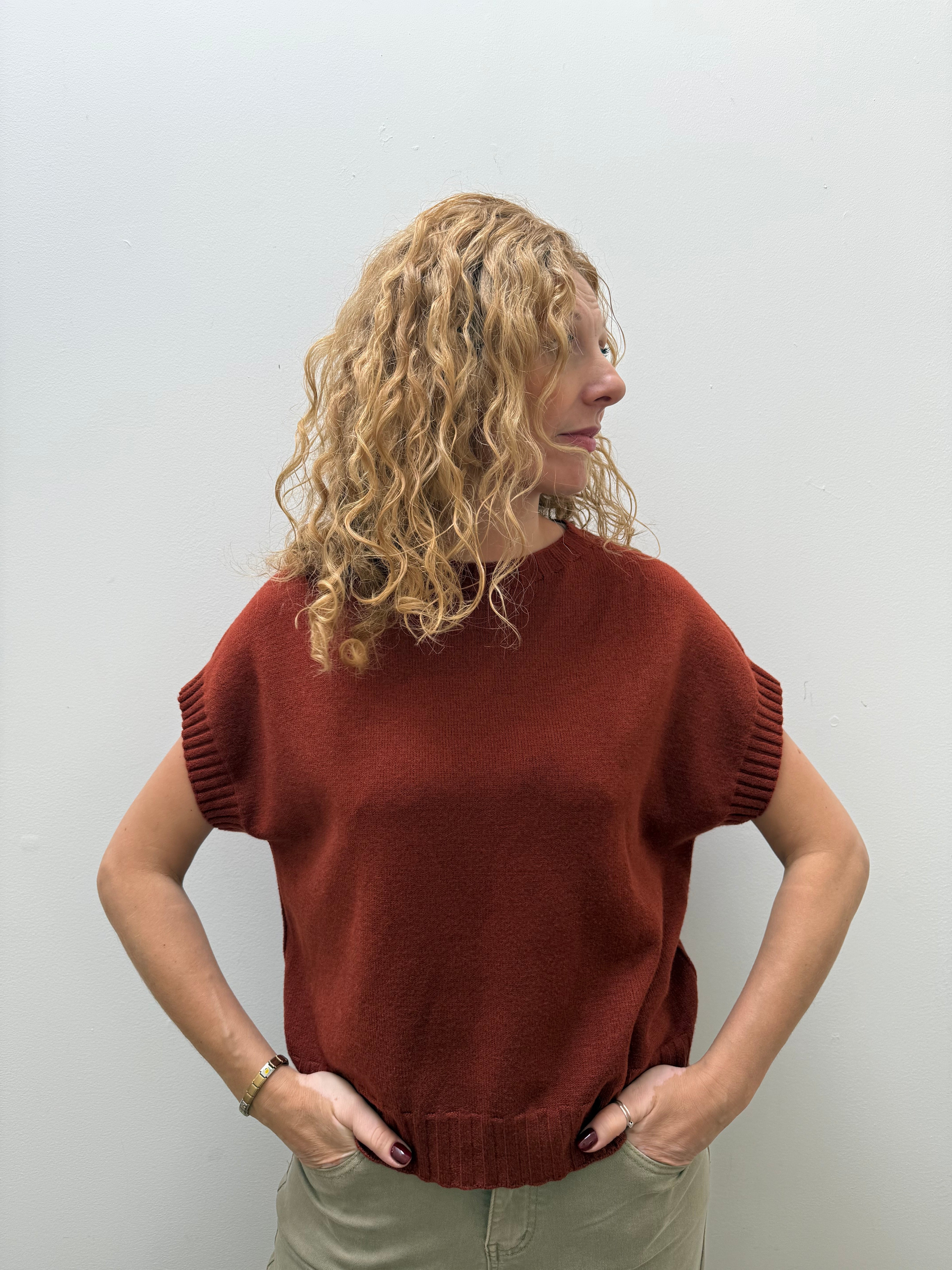 A woman with curly blonde hair wearing a rust-colored short-sleeve knit sweater and khaki pants, standing against a plain light gray wall.