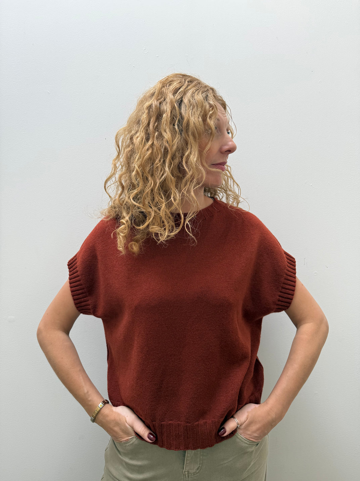 A woman with curly blonde hair wearing a rust-colored short-sleeve knit sweater and khaki pants, standing against a plain light gray wall.