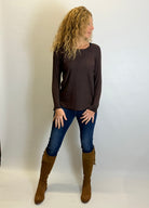 Woman wearing a brown long-sleeve top, blue jeans, and brown boots against a white background