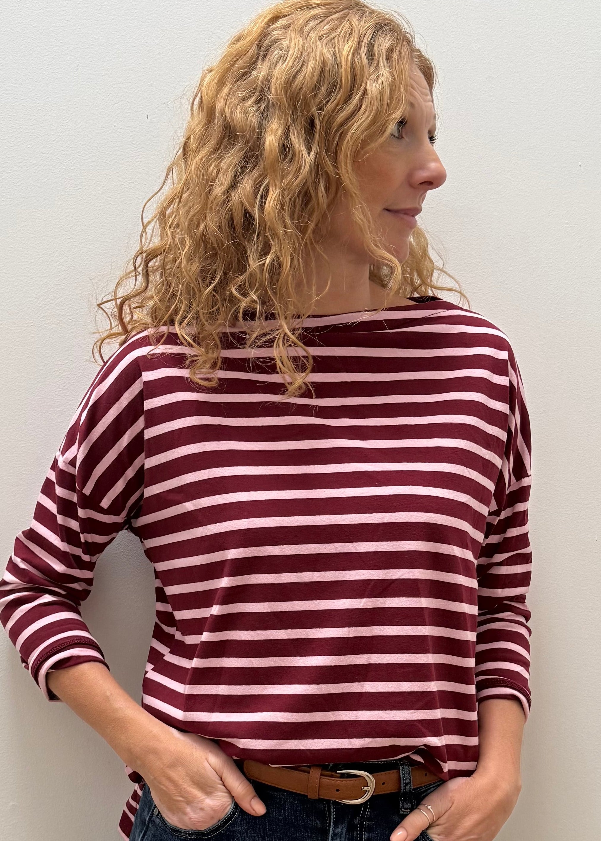 A woman with curly blonde hair wearing a burgundy and white striped long-sleeve top, dark blue jeans, and a brown belt, standing against a plain off-white wall.