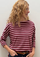 A woman with curly blonde hair wearing a burgundy and white striped long-sleeve top, dark blue jeans, and a brown belt, standing against a plain off-white wall.