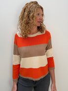 A woman with curly blonde hair wearing a horizontally striped knit sweater in orange, beige, and cream, paired with dark gray pants, standing against a plain white wall.