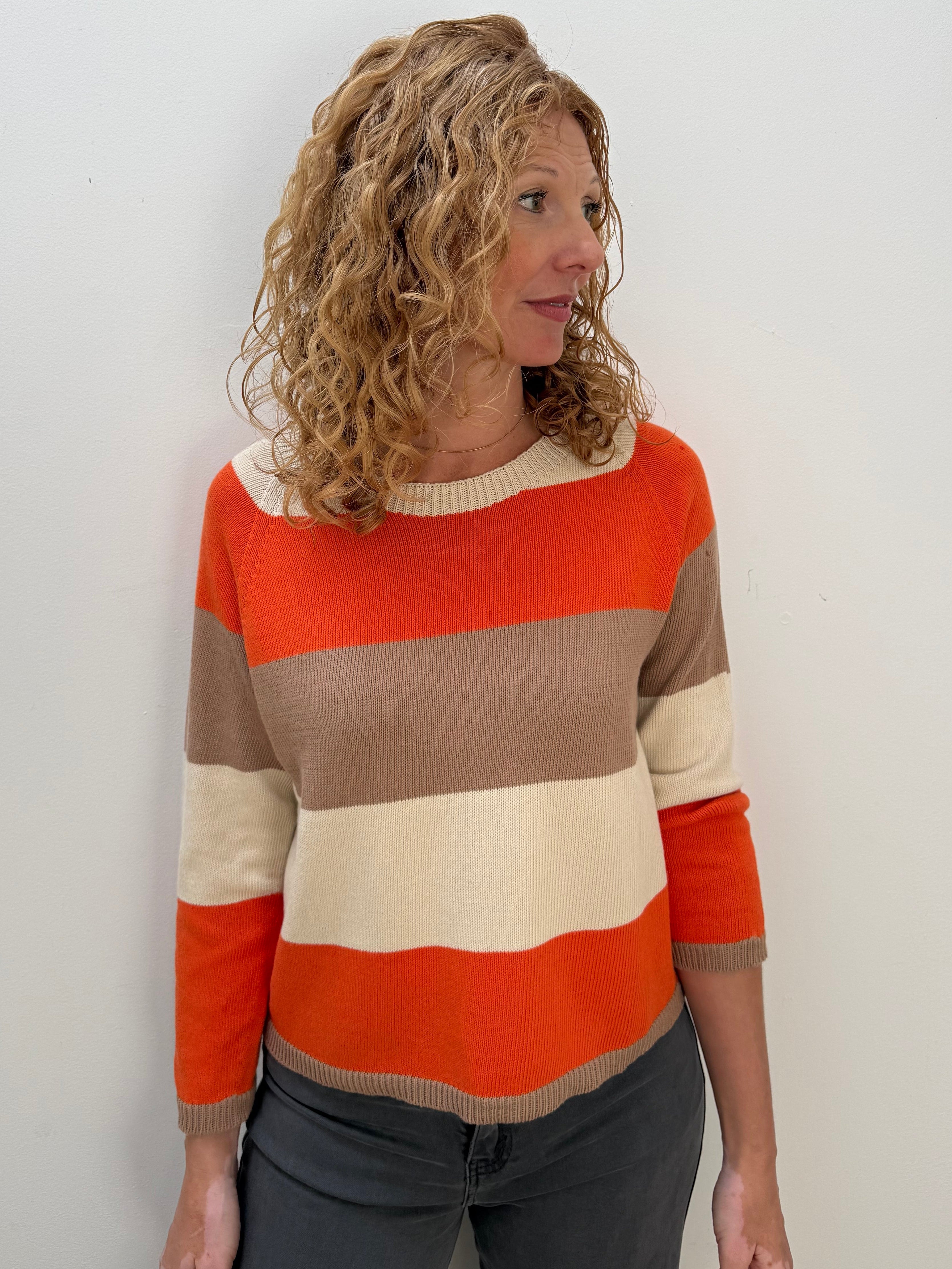 A woman with curly blonde hair wearing a horizontally striped knit sweater in orange, beige, and cream, paired with dark gray pants, standing against a plain white wall.